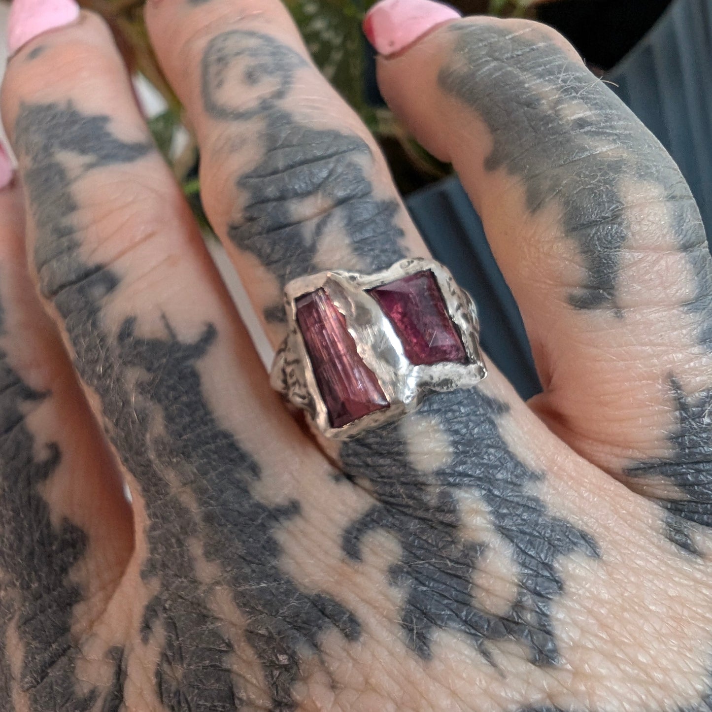 Hand with tattoos wearing a ring with two pink gemstones against a blurred background